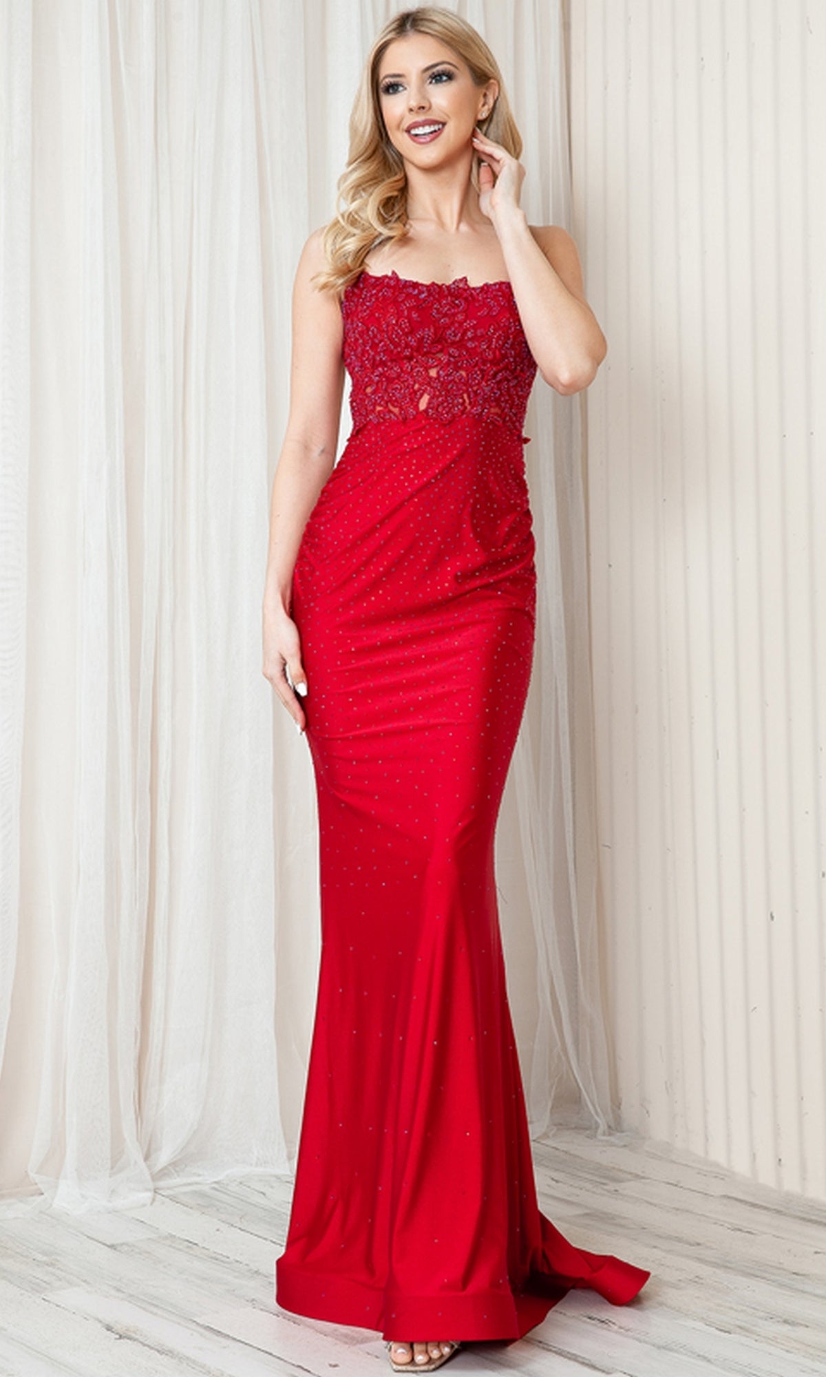 A woman stands indoors against a light curtain and wall, wearing the Long Beaded Prom Dress with Lace Bodice TM1001—a sleeveless, floor-length red gown featuring a sheer lace bodice.